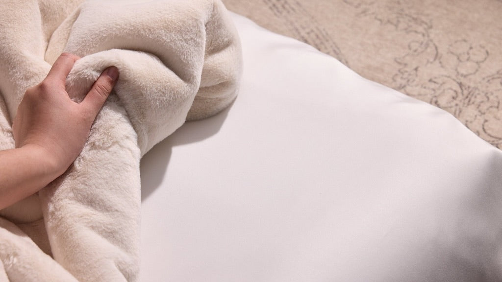 A close-up of a hand pulling back a soft cream-colored plush blanket to reveal a smooth white pillow underneath, with a patterned beige bedspread in the background.